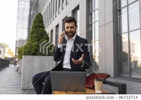 Smiling busy young european man manager with beard in suit with cup of coffee and laptop calls by phone Smiling busy young european man manager with beard in suit with cup of coffee and laptop calls by phone 96733714