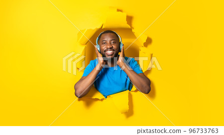 Happy african american man wearing wireless headphones and enjoying music, posing through hole in torn yellow paper 96733763
