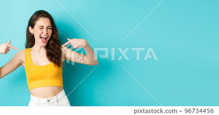 I am the best. Smiling confident woman feeling self-assured, pointing at herself and winking happy, self-promoting own abilities, standing against blue background 96734456