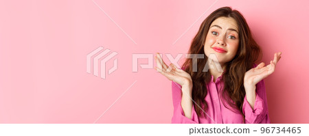 Close up portrait of clueless cute woman, shrugging shoulders and smiling with unaware face expression, standing over pink background 96734465