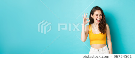 Young cheerful lady in summer clothes winking and saying yes, alright gesture, making OK sign in approval, standing against blue background 96734561