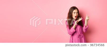 Annoyed girlfriend arguing about proposal, wants to get married, showing finger without engagement ring, standing confused against pink background 96734566