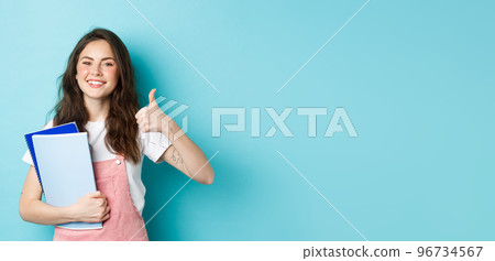 Young woman attend courses, girl student studying, holding notebooks and showing thumb up in approval, recommending company, standing over blue background Young woman attend courses, girl student studying, holding notebooks and showing thumb up in approval, recommending company, standing over blue background 96734567