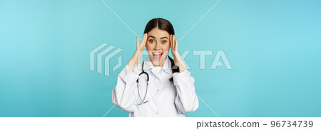 Portrait of surprised and excited smiling doctor woman, holding hands on head, reaction to mindblowing awesome news, standing over blue background Portrait of surprised and excited smiling doctor woman, holding hands on head, reaction to mindblowing awesome news, standing over blue background 96734739