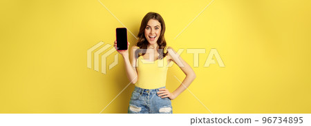 Stylish smiling woman showing mobile phone screen, app interface on smartphone, standing in tank top and shorts against yellow background Stylish smiling woman showing mobile phone screen, app interface on smartphone, standing in tank top and shorts against yellow background 96734895