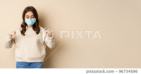 Cute girl in medical face mask, pointing fingers and looking down, showing advertisement below, standing against beige background 96734896