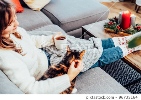 Relaxed woman with cup of hot drink petting cat on the sofa at home while watching movie, TV with christmas decoration atmosphere. Lady wear jumper and warm socks. Cozy and comfortable winter concept Relaxed woman with cup of hot drink petting cat on the sofa at home while watching movie, TV with christmas decoration atmosphere. Lady wear jumper and warm socks. Cozy and comfortable winter concept 96735164