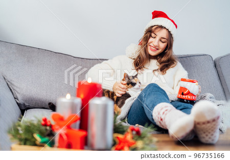 Woman in santa hat with tea cup and plaid petting pet cat on the sofa at home while watching movie, TV with christmas decoration atmosphere. Girl wear warm socks. Cozy and comfortable winter concept 96735166