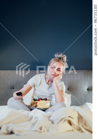 Bored, sad woman in home clothes eating cake and using the remote control to change the channel while watching television in bed at night. Overeating, Jamming of negative emotions. Vertical card. Bored, sad woman in home clothes eating cake and using the remote control to change the channel while watching television in bed at night. Overeating, Jamming of negative emotions. Vertical card. 96735180