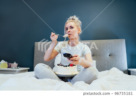 Surprised, stressed, shocked woman eating cake and holding a remote control while watching TV in bed. Scary horror movie or terrible news, scare in her eyes Overeating, Jamming of negative emotions 96735181