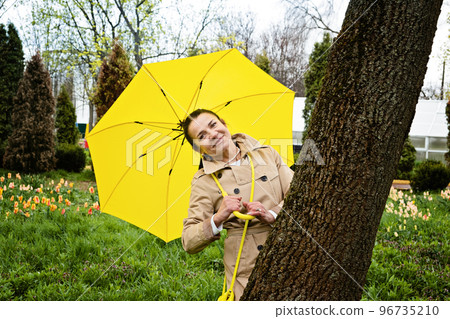 Happy senior woman in yellow rain coat with yellow umbrella walking in park. Cheerful mature, elderly, retired Latina Hispanic woman enjoying life at rainy day outdoors 96735210
