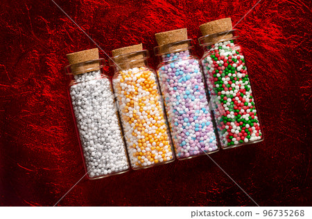 Nonpareils in small glass bottles, on a dark red and surface. Winter and christmas colored mixes of edible confectionery of tiny sugar balls. Hundreds and Thousands, used as decoration and as topping. 96735268