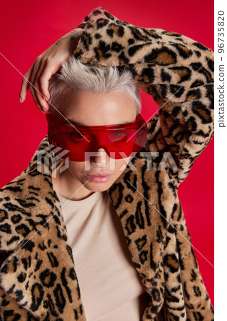 Portrait of young stylish woman with short hair posing in stylish coat with animal print and glasses isolated over red background 96735820