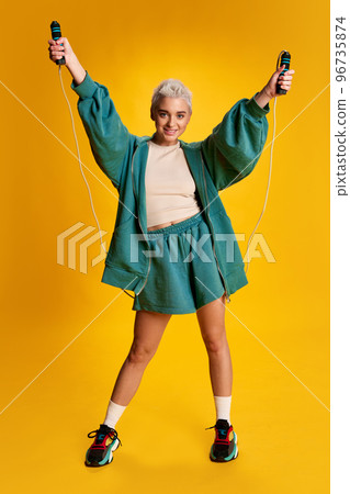 Portrait of young stylish woman with short hair posing comfy sportswear and jumping rope isolated over yellow background 96735874