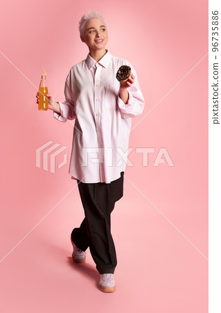Portrait of young stylish woman with short hair posing with donut and orange juice isolated over pink background 96735886