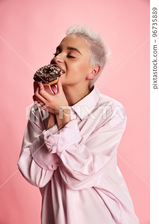Portrait of young stylish woman with short hair posing, eating donut isolated over pink background. Dessert Portrait of young stylish woman with short hair posing, eating donut isolated over pink background. Dessert 96735889
