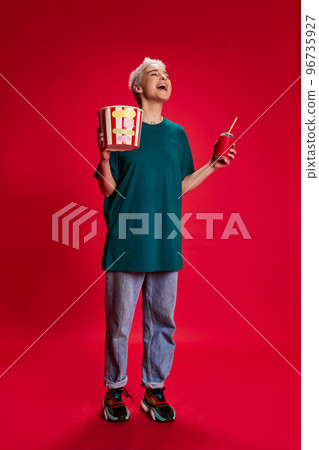 Portrait of young stylish woman with short hair posing with popcorn basket and laughing isolated over red background. Comedy 96735927