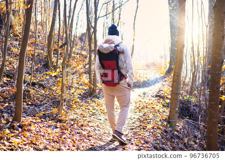 Sporty father carrying his infant son wearing winter jumpsuit and cap in backpack carrier hiking in autumn forest 96736705