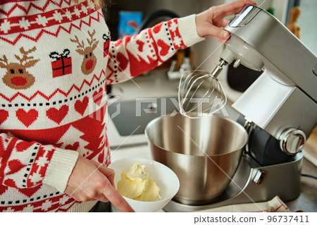 Woman in festive christmas sweater preparing dough for cookies at home kitchen. Female hands use electric mixer. Modern kitchen household appliances Woman in festive christmas sweater preparing dough for cookies at home kitchen. Female hands use electric mixer. Modern kitchen household appliances 96737411
