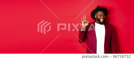 Christmas shopping and people concept. Handsome Black man showing ok sign with confidence, give guarantee, saying yes, standing over red background Christmas shopping and people concept. Handsome Black man showing ok sign with confidence, give guarantee, saying yes, standing over red background 96738732