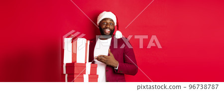 Christmas, New Year and shopping concept. Cheerful Black man secret santa holding xmas presents and smiling excited, bring gifts, standing against red background 96738787