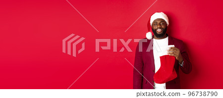 Handsome african american man celebrating winter holidays, holding Christmas sock and smiling, wearing santa hat, standing over red background 96738790