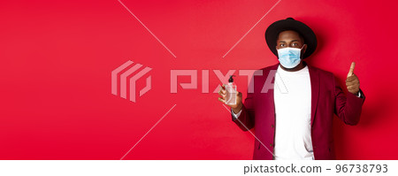 Covid-19, quarantine and holidays concept. Young african american man in medical mask, showing hand sanitizer and thumb up, recommending to use antiseptic Covid-19, quarantine and holidays concept. Young african american man in medical mask, showing hand sanitizer and thumb up, recommending to use antiseptic 96738793
