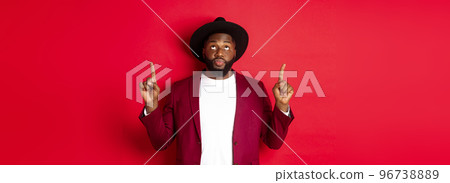 Winter holidays and shopping concept. Curious african american guy in party clothing looking and pointing fingers up, reading promo sign, standing over red background 96738889