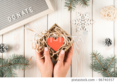 Female hands hold present donation box with red paper heart postcard, top view. Decorative spruce branches with cones and snowflake on white planking. Giving money. Christmas or New Year charity 96740357