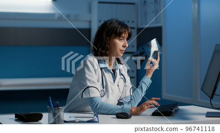 Practitioner holding radiography analyzing lungs before writing medical expertise on computer, planning health care treatment to cure patient disease. Medic working over hours in hospital office 96741118