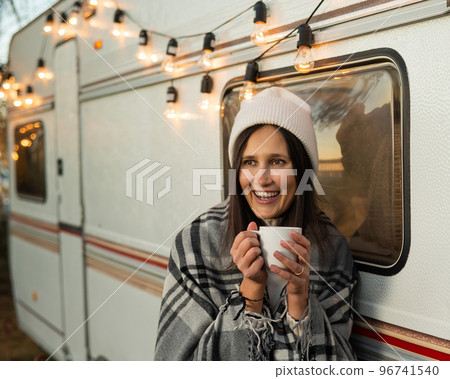 Caucasian woman in a knitted hat wrapped in a plaid and drinks a warming drink outdoors. Travel in a motor home in the fall. 96741540