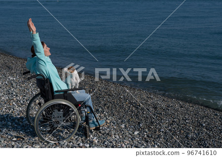 Happy woman in a wheelchair on the seashore raised her hands and imagines that she is flying.  96741610