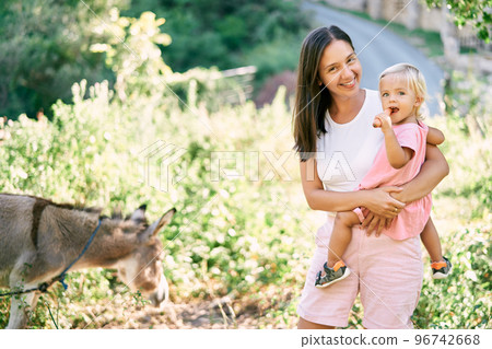 Smiling mother with a little girl chewing a carrot stands in a pasture next to a donkey. High quality photo Smiling mother with a little girl chewing a carrot stands in a pasture next to a donkey. High quality photo 96742668