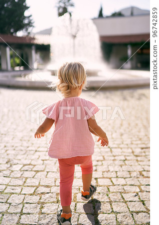 Little girl walks along the paving stones in the square towards the fountain. Back view. High quality photo 96742859