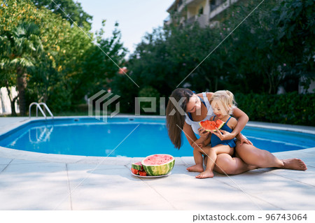 Mom feeds watermelon to a little girl sitting on her lap near the pool. High quality photo Mom feeds watermelon to a little girl sitting on her lap near the pool. High quality photo 96743064