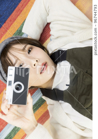 Young woman lying on a rug and holding a film camera Young woman lying on a rug and holding a film camera 96744793