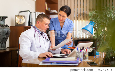 Two fellow doctors filling up medical forms on laptop in office Two fellow doctors filling up medical forms on laptop in office 96747303