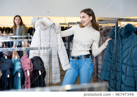 Female purchaser choosing winter down jacket in shopping center Female purchaser choosing winter down jacket in shopping center 96747349