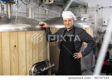 Adult happy brewer is standing near reservoir for beer in fabric Adult happy brewer is standing near reservoir for beer in fabric 96747384