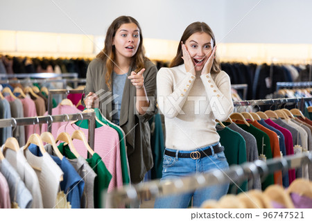 Amazed female holding warm clothes on hangers and showing her friend a discount during shopping in a store Amazed female holding warm clothes on hangers and showing her friend a discount during shopping in a store 96747521