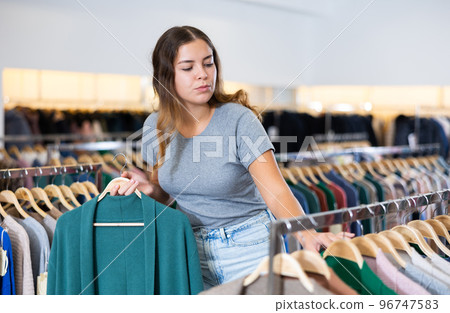 Serious young woman holding a turquoise cardigan on a rack while choosing clothes in a boutique Serious young woman holding a turquoise cardigan on a rack while choosing clothes in a boutique 96747583