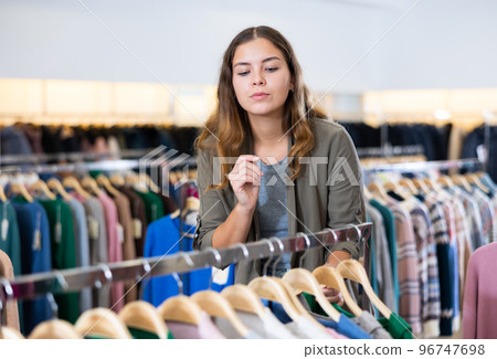 Positive young woman choosing season outfit clothes among the racks in a shopping mall Positive young woman choosing season outfit clothes among the racks in a shopping mall 96747698