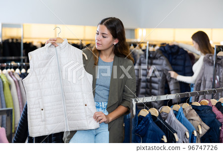 Young casual female keeping white waistcoat on a rack while shopping in the in a mall Young casual female keeping white waistcoat on a rack while shopping in the in a mall 96747726