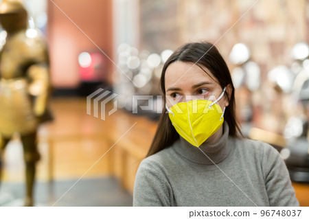 Female museum visitor in protective mask examining ancient armor 96748037