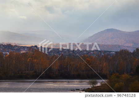 Autumn landscape of Eastern Siberia. The city on the banks of the river is illuminated by the rays of the setting sun. The sun shines through the clouds. Fog in the mountains. 96748128