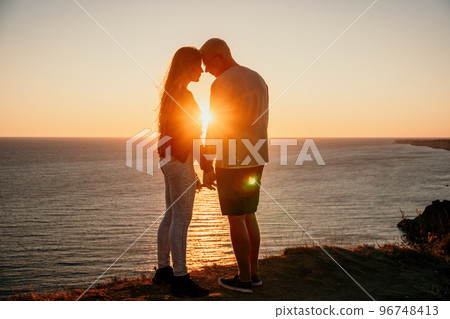 Silhouette of a young romantic couple enjoying an evening on a cliff above the sea with a red burning sunset. Traveling couple, man and woman, holding hands and standing on top of a mountain at sunset 96748413