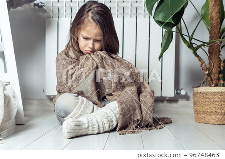 Depressed, cried, serious little girl wrapped in warm plaid, trying warming up, keeping warm, sit near heating radiator Depressed, cried, serious little girl wrapped in warm plaid, trying warming up, keeping warm, sit near heating radiator 96748463