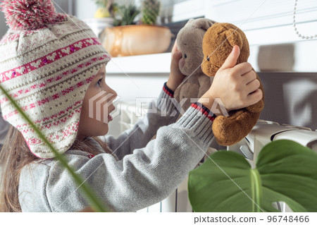 Close up calm concentrated little girl in knitted hat, sweater warm up with best friends stuffed toys near heat radiator Close up calm concentrated little girl in knitted hat, sweater warm up with best friends stuffed toys near heat radiator 96748466
