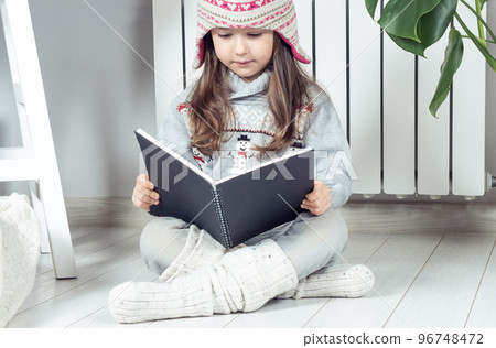 Pensive serious cute little girl in wool hat and warm clothes with snowman reading book or notepad near heating radiator Pensive serious cute little girl in wool hat and warm clothes with snowman reading book or notepad near heating radiator 96748472