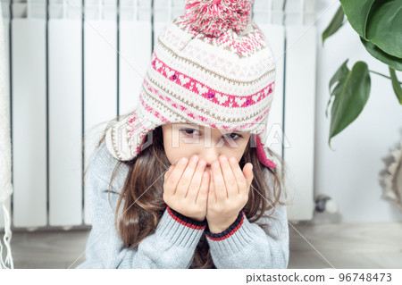 Close up little brunette girl face breathing on hands to keep warm near cold broken heating radiator, looking at camera Close up little brunette girl face breathing on hands to keep warm near cold broken heating radiator, looking at camera 96748473